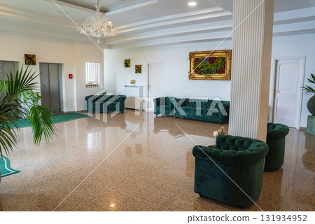 Interior of a hotel lobby in Amalfi, Italy, featuring marble flooring, green velvet seating, a chandelier, gold framed artwork, and decorative plants. 131934952