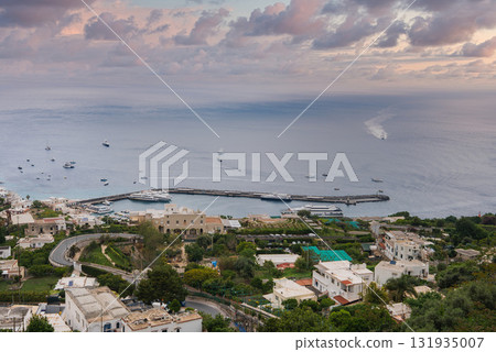 The island of Capri in Italy features a harbor with yachts, whitewashed buildings, lush greenery, winding roads, and a curved pier under a colorful sky. 131935007