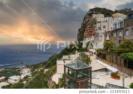 The island of Capri in Italy at sunset, featuring colorful cliffside buildings, a glass elevator, the Mediterranean Sea, boats, and lush greenery. 131935011