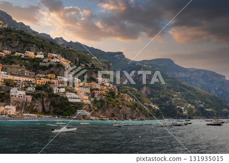 Positano's vibrant buildings cascade down a steep cliff, surrounded by greenery, mountains, and the Mediterranean Sea under a dramatic cloudy sky. 131935015