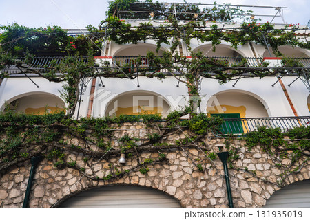 A multi level building in Positano, Italy, featuring arched balconies with greenery, flowering vines, and rustic stonework in a coastal Mediterranean style. A multi level building in Positano, Italy, featuring arched balconies with greenery, flowering vines, and rustic stonework in a coastal Mediterranean style. 131935019