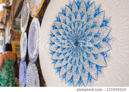 A detailed ceramic plate with a blue and white floral design is displayed outdoors in Ravello, Italy, with other plates and a passerby in view. A detailed ceramic plate with a blue and white floral design is displayed outdoors in Ravello, Italy, with other plates and a passerby in view. 131935024