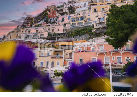 Positano's pastel colored buildings rise vertically on steep terrain, framed by flowers and a tree, under a pink and purple sunset sky. 131935042