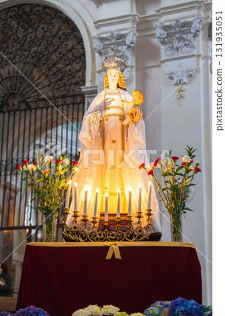 Statue of Virgin Mary with infant Jesus on a red velvet pedestal, surrounded by candles and flowers, set against a wrought iron gate in Ravello, Italy. Statue of Virgin Mary with infant Jesus on a red velvet pedestal, surrounded by candles and flowers, set against a wrought iron gate in Ravello, Italy. 131935051