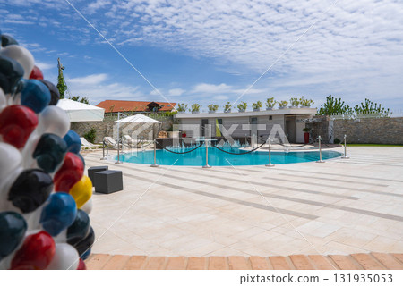 Outdoor pool with tiled deck, white umbrellas, lounge chairs, and a colorful sculpture. Modern building, stone wall, and greenery in the background. Outdoor pool with tiled deck, white umbrellas, lounge chairs, and a colorful sculpture. Modern building, stone wall, and greenery in the background. 131935053