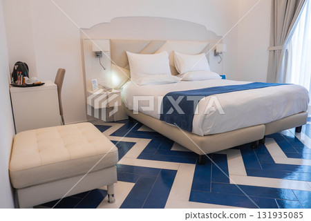 Bright hotel room featuring a double bed with white linens, blue runner, chevron patterned tiled floor, desk, coffee station, and natural light. 131935085