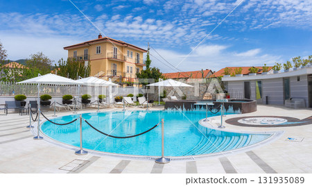 Circular pool with white lounge chairs and umbrellas, bordered by a tiled deck. Yellow multi story building and greenery under a blue sky in Amalfi, Italy. Circular pool with white lounge chairs and umbrellas, bordered by a tiled deck. Yellow multi story building and greenery under a blue sky in Amalfi, Italy. 131935089