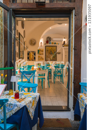 The image shows a restaurant entrance in Amalfi, Italy, with turquoise chairs, white tables, colorful decor, ceramic plates, and arched ceilings. 131935097