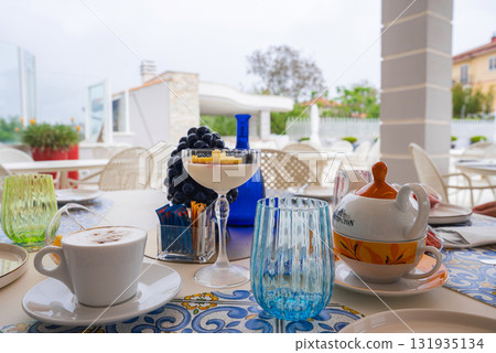 A vibrant outdoor dining table in Amalfi, Italy, featuring a cappuccino, dessert, floral teapot, colorful glassware, and lush greenery in the background. A vibrant outdoor dining table in Amalfi, Italy, featuring a cappuccino, dessert, floral teapot, colorful glassware, and lush greenery in the background. 131935134