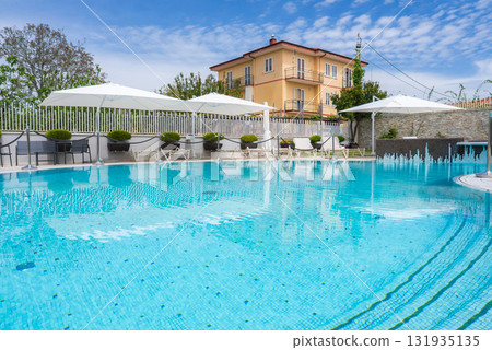 Serene pool with clear water, white lounge chairs, and umbrellas. Yellow villa with balconies and terracotta roof in Amalfi, Italy under a blue sky. Serene pool with clear water, white lounge chairs, and umbrellas. Yellow villa with balconies and terracotta roof in Amalfi, Italy under a blue sky. 131935135