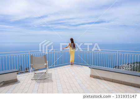 A woman in a yellow dress stands on a tiled terrace with a white railing, overlooking the Tyrrhenian Sea, coastline, and distant buildings in Amalfi. A woman in a yellow dress stands on a tiled terrace with a white railing, overlooking the Tyrrhenian Sea, coastline, and distant buildings in Amalfi. 131935147