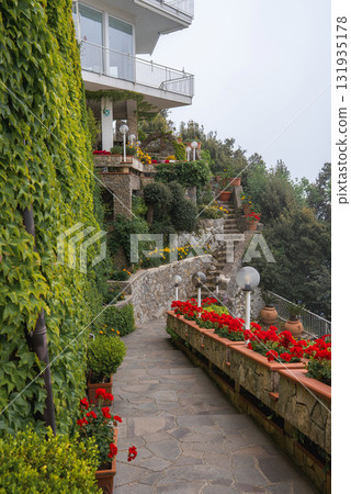 A stone pathway lined with red flowers in terracotta pots leads to a staircase near a luxury building with ivy covered balconies in Amalfi, Italy. 131935178