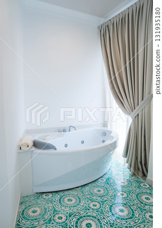 White jacuzzi style bathtub with built in jets, turquoise and white patterned tiles, and natural light through a tied back beige curtain in Amalfi. White jacuzzi style bathtub with built in jets, turquoise and white patterned tiles, and natural light through a tied back beige curtain in Amalfi. 131935180