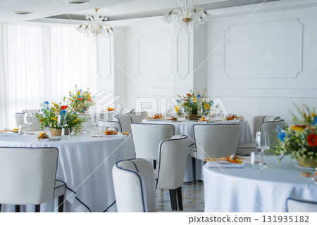 Refined dining space with white walls, ornate chandeliers, floral centerpieces, and natural light, likely in a luxury venue in Amalfi, Italy. 131935182