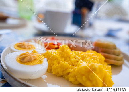 A detailed view of a breakfast plate with scrambled eggs, halved boiled eggs, smoked salmon, and sliced vegetables. A coffee cup is blurred in the background. A detailed view of a breakfast plate with scrambled eggs, halved boiled eggs, smoked salmon, and sliced vegetables. A coffee cup is blurred in the background. 131935183
