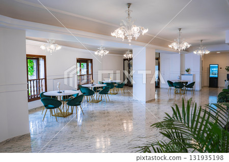 Bright interior with round tables, green chairs, marble flooring, modern chandeliers, large windows, and decorative plants in Amalfi, Italy. 131935198