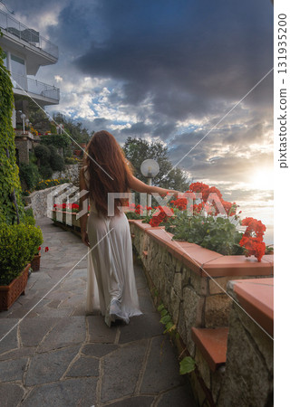 A woman with red hair walks on a stone paved terrace in Amalfi, Italy, surrounded by red flowers, lush greenery, and a view of the Tyrrhenian Sea. A woman with red hair walks on a stone paved terrace in Amalfi, Italy, surrounded by red flowers, lush greenery, and a view of the Tyrrhenian Sea. 131935200