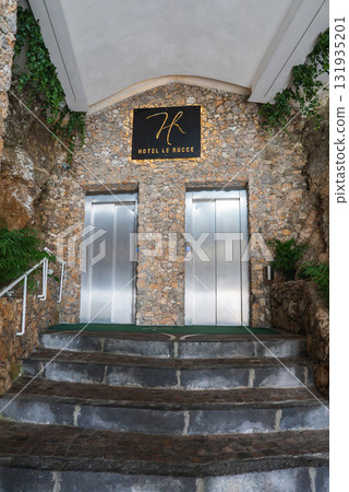 Modern stainless steel elevators set in a rustic stone wall, framed by greenery, with a curved stone staircase leading to Hotel Le Rocce in Amalfi, Italy. Modern stainless steel elevators set in a rustic stone wall, framed by greenery, with a curved stone staircase leading to Hotel Le Rocce in Amalfi, Italy. 131935201