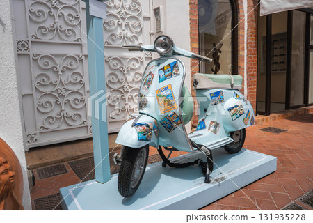A light blue scooter adorned with Capri themed stickers and postcards stands before a white wrought iron gate and brick framed storefront in Capri, Italy. 131935228