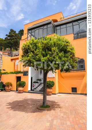 Vibrant orange building with large glass windows, a tree in the courtyard, terracotta pots, stone walls, and lush greenery on Capri Island, Italy. 131935233