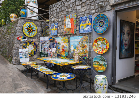 A vibrant ceramic shop in Ravello, Italy, featuring colorful plates, tiles, and tables with coastal and floral designs against a stone wall backdrop. 131935246
