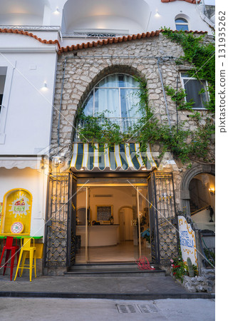 The stone facade of Hotel Reginella features an arched window, ivy covered walls, a striped awning, and a cafe area with colorful stools in Positano, Italy. 131935262