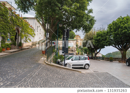 Charming street in Ravello, Italy, with cobblestone pavement, ivy covered building, parked cars, leafy trees, and hilly landscape under a cloudy sky. 131935263