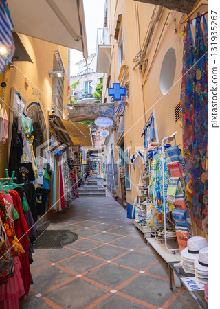 Narrow street in Positano, Italy, lined with shops displaying clothing and souvenirs. Warm toned buildings and a pharmacy sign add unique details. 131935267