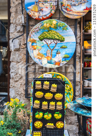 Ceramic plates with coastal scenes and lemon shaped magnets displayed against a stone wall in Ravello, Italy, highlighting local craftsmanship. 131935269