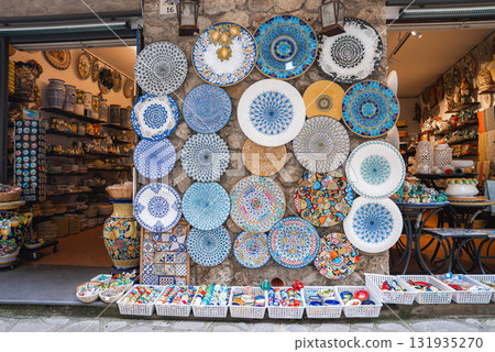 Vibrant ceramic shop in Ravello, Italy, featuring colorful plates on a stone wall, baskets of ornaments, and a glimpse of the artistic interior. 131935270