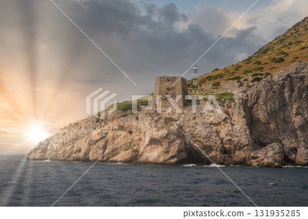 A coastal cliff in Positano, Italy, features a historic stone tower and a lighthouse. The setting sun casts warm light over the sea and dramatic clouds. 131935285