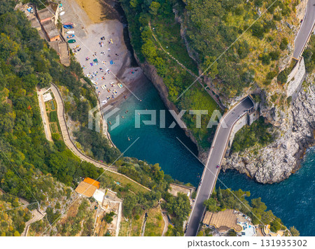 Aerial image of a cove on the Amalfi Coast with turquoise waters, steep cliffs, a beach with umbrellas, a bridge, terraced gardens, and lush greenery. 131935302