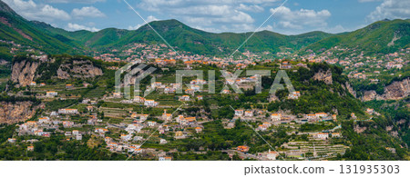 Aerial view of the Amalfi region in Italy, showcasing terraced hills, pastel colored buildings, rugged cliffs, and rolling green mountains under a cloudy sky. 131935303