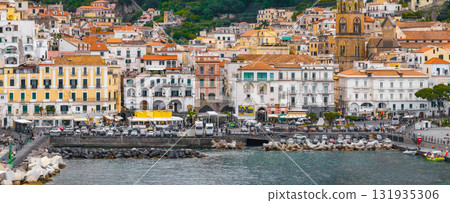 View of Amalfi town with pastel colored hillside buildings, the Amalfi Cathedral bell tower, a busy waterfront, and lush green hills in the background. 131935306