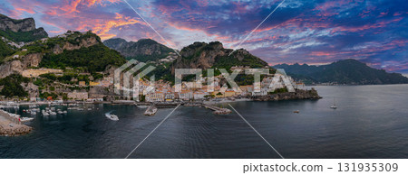 Aerial view of Amalfi, Italy, featuring colorful buildings on cliffs, a hillside hotel, a harbor with boats, and a vibrant pink and purple sunset sky. 131935309