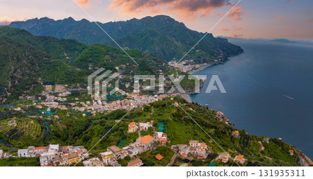 Aerial view of Ravello, Italy, with terraced hillsides, orange tiled roofs, winding roads, and the Amalfi Coast meeting the calm blue sea at sunset. 131935311