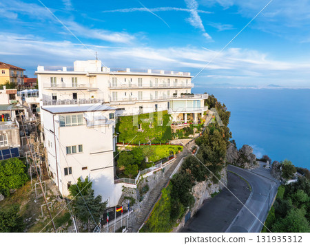 Luxurious white hotel with green facade on a cliff in Amalfi, Italy. Terraces face the sea, with a winding road and trees under a blue sky with clouds. 131935312