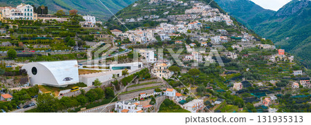 Aerial view of Ravello, Italy, featuring terraced gardens, pastel buildings, a modern curved roof structure, and verdant mountains in the background. 131935313