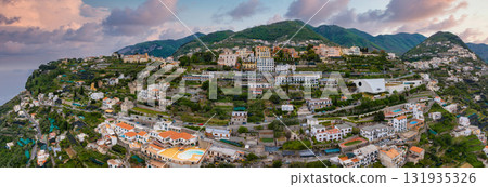 Aerial view of Ravello, Italy, with terraced greenery, red tiled roofs, a modern white building, and mountains leading to the coastline at sunset. 131935326