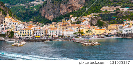 Aerial view of Amalfi, Italy, showing colorful hillside buildings, a domed church, dramatic cliffs, a pier, and the vibrant blue Tyrrhenian Sea. 131935327