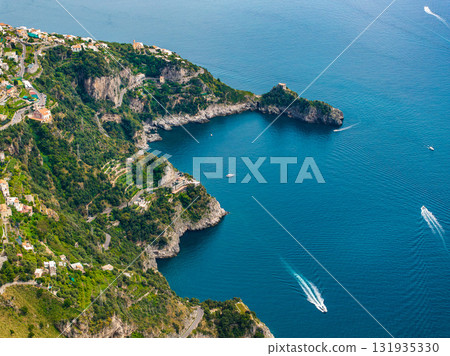 Aerial view of the Amalfi Coast in Italy, showing cliffs, green hills, a small bay, boats on the Tyrrhenian Sea, and villas along winding roads. 131935330