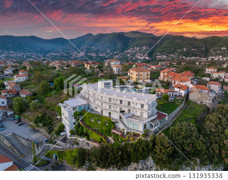 Aerial view of a luxury hotel in Amalfi, Italy, with terraces, a rooftop pool, red tiled houses, lush greenery, and a vibrant sunset sky. 131935338