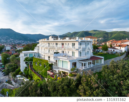 A multi story white building with balconies and rooftop area on a hillside, surrounded by greenery, with red roofed town and hills in the distance. A multi story white building with balconies and rooftop area on a hillside, surrounded by greenery, with red roofed town and hills in the distance. 131935382