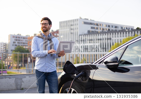 A man stands next to his electric vehicle while using a laptop, possibly monitoring the charging process. A man stands next to his electric vehicle while using a laptop, possibly monitoring the charging process. 131935386