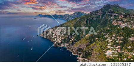 Aerial view of Amalfi, Italy, showing cliffs, green hills, whitewashed buildings, a luxury hotel, boats on the sea, and a vibrant sunset sky. Aerial view of Amalfi, Italy, showing cliffs, green hills, whitewashed buildings, a luxury hotel, boats on the sea, and a vibrant sunset sky. 131935387