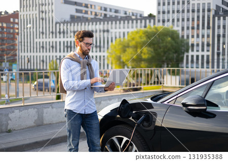 A man uses a laptop while his electric vehicle charges, showcasing modern technology and sustainable transportation in an urban setting. A man uses a laptop while his electric vehicle charges, showcasing modern technology and sustainable transportation in an urban setting. 131935388