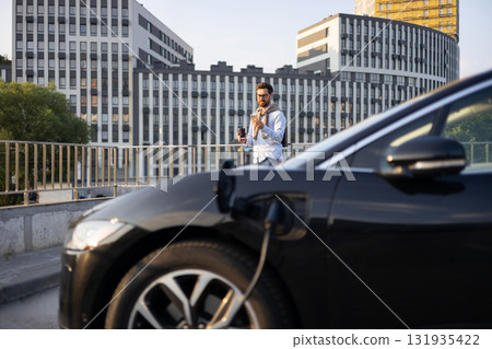 A man checks his phone while his electric vehicle charges in front of modern buildings. 131935422