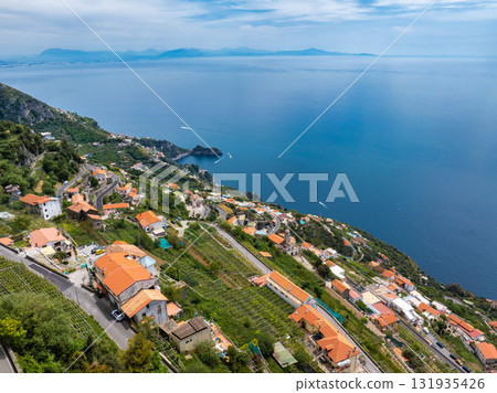 Aerial view of Amalfi, Italy, featuring orange roofed buildings on terraced hills, lush greenery, a winding road, and the expansive Tyrrhenian Sea. 131935426