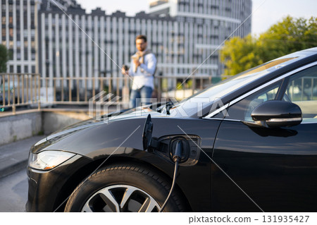 A black electric vehicle is charging, with a man standing in the background. A black electric vehicle is charging, with a man standing in the background. 131935427