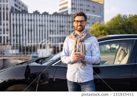 A man smiles while holding his phone next to his electric vehicle while it charges in an urban setting. 131935431